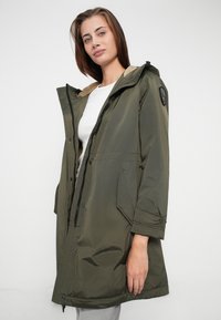 Olīvu zaļš parka jakas ar rāvējslēdzēju un pogām, kas ietver kapuci, sānu kabatas un mīkstu iekšējo oderi. Strukturēts, viegls audums.