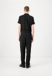 Chemise noire à manches courtes et pantalon assorti en tissu texturé, coupe droite, avec poches arrière, accompagnés de chaussures noires.