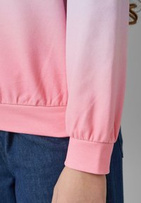 Felpa rosa ombre con una texture morbida, dotata di polsini arricciati e orlo a costine di una tonalità di rosa più scura. Indossata su un paio di jeans.