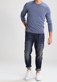 Maglione blu a maglia con scollatura rotonda, abbinato a jeans in denim scuro e sneakers bianche. I jeans sono leggermente arrotolati sui polsini.