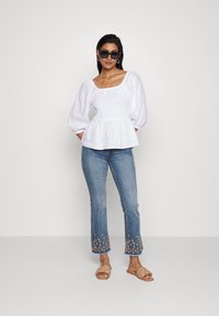 Top peplum branco com decote quadrado e mangas bufantes, combinado com jeans azuis de boca de sino com bordados florais na bainha e sandálias bege.