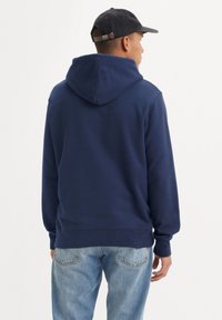 Marineblauwe hoodie met een ruime capuchon, geribbelde mouwen en tailleband, gemaakt van zachte stof. Draagt met lichtblauwe jeans en een donkere pet.