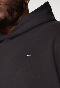 Juoda hoodie, pagaminta iš minkštos medžiagos, su V formos iškirptė ir maža, spalvota logotipo emblema ant krūtinės. Minkšta tekstūra.