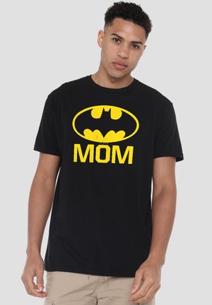 Junger Mann trägt ein schwarzes T-Shirt mit gelbem Batman-Logo und dem Wort "MOM" in großen Buchstaben, steht vor einem einfarbigen Hintergrund.