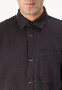 Camicia button-up marrone scuro con colletto a punta, tasca anteriore e bottoni in plastica nera. Il tessuto appare testurizzato e robusto.