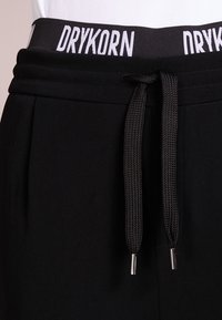 Czarne spodnie dresowe z szerokim, elastycznym pasem z białym napisem "DRYKORN" oraz sznurkiem do regulacji dopasowania. Miękka faktura tkaniny.