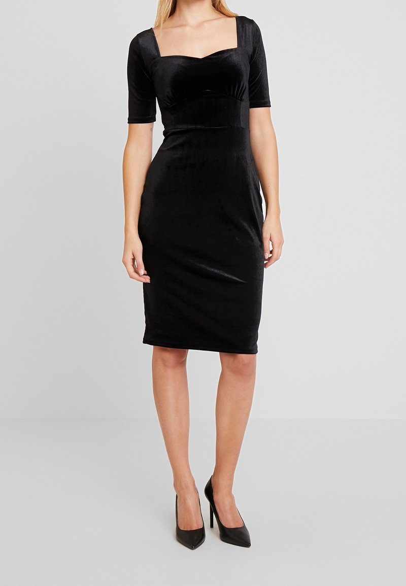 Femme portant une robe noire cintrée longueur genou avec manches courtes et encolure carrée, associée à des talons noirs, debout sur un fond gris.