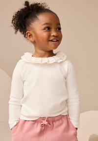 Bambino con i capelli ricci, indossa un top bianco con colletto arricciato e pantaloni rosa, sorride e guarda di lato, su uno sfondo beige.