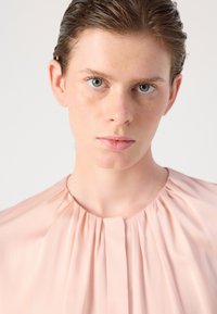 Blusa rosa chiaro con design plissettato, scollatura rotonda e texture di tessuto morbido. Inquadratura ravvicinata del capo indossato da un modello.