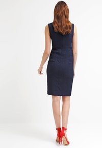 Marineblauwe mouwloze jurk met een aangesloten silhouette, knielang, textiel met structuur en een gladde rugnaad. Gecombineerd met rode high heels.