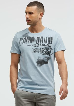 Man with short hair wearing light blue "CAMP DAVID" printed t-shirt and gray pants, looking left with one hand in pocket.