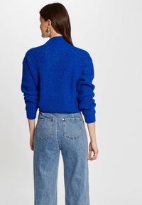 Pull en tricot bleu avec une texture côtelée, col montant et épaules tombantes, assorti à un jean en denim bleu clair taille haute.