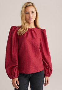 Blusa rossa con maniche a sbuffo, colletto rotondo, tessuto texturizzato e dettagli arricciati ai polsini delle maniche, abbinata a jeans scuri.