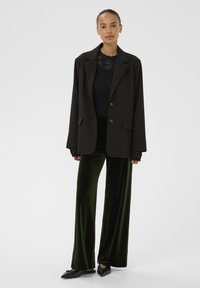 Blazer negru cu buzunare aplicate peste un top din dantelă neagră, asociat cu pantaloni largi din catifea verde închis și pantofi negri cu vârf ascuțit.