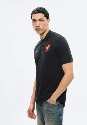 Młody mężczyzna z krótkimi kręconymi włosami, ubrany w czarną koszulkę polo z pomarańczowym herbem lwa i dżinsy, stojący z jedną ręką w kieszeni.