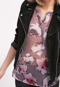 Giacca in suede nera sopra una blusa trasparente a fiori con motivi di fiori rosa e bianchi, scollo a V e tessuto leggero e fluido.