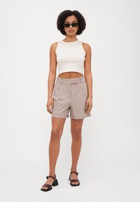 Top scurt de culoare bej deschis, asortat cu pantaloni scurți gri, cu talie înaltă și croi elegant. Modelul poartă ochelari de soare mari și negri și sandale cu toc.