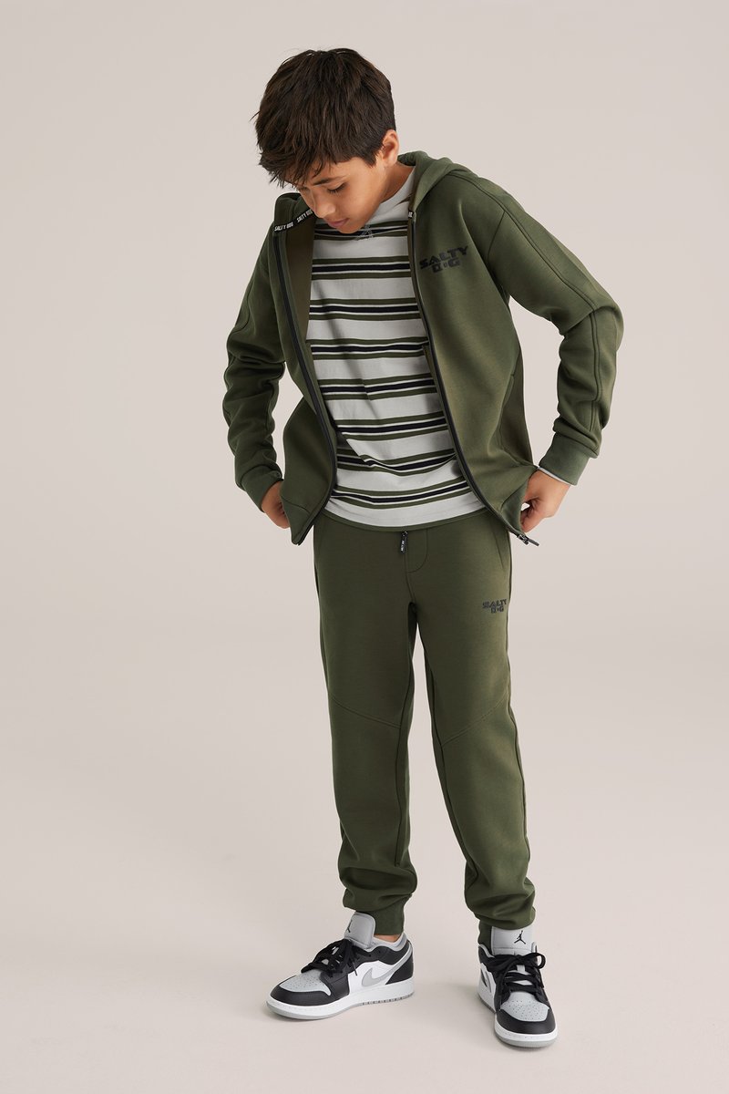 Zelená mikina a zodpovedajúce teplákové nohavice s čiernym logom, vrstvené cez pruhované tričko. Oblečenie dopĺňajú tenisky s čierno-bielymi akcentmi.