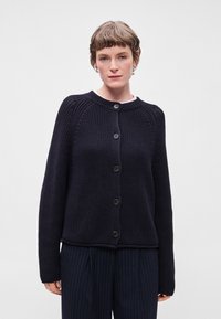 Σκούρο ναυτικό πλεκτό ζακέτα με στρογγυλή λαιμόκοψη, πέντε μεγάλες κουμπόθηκες και μακριά μανίκια. Ριγωτή υφή και ελαφρώς κομμένη εφαρμογή.