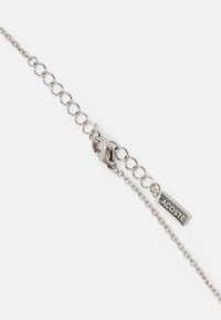 Sølv metal kædearmbånd med ovale led og en lille klipslås. Har et vedhæng med "LACOSTE" graveret. Minimalistisk design.