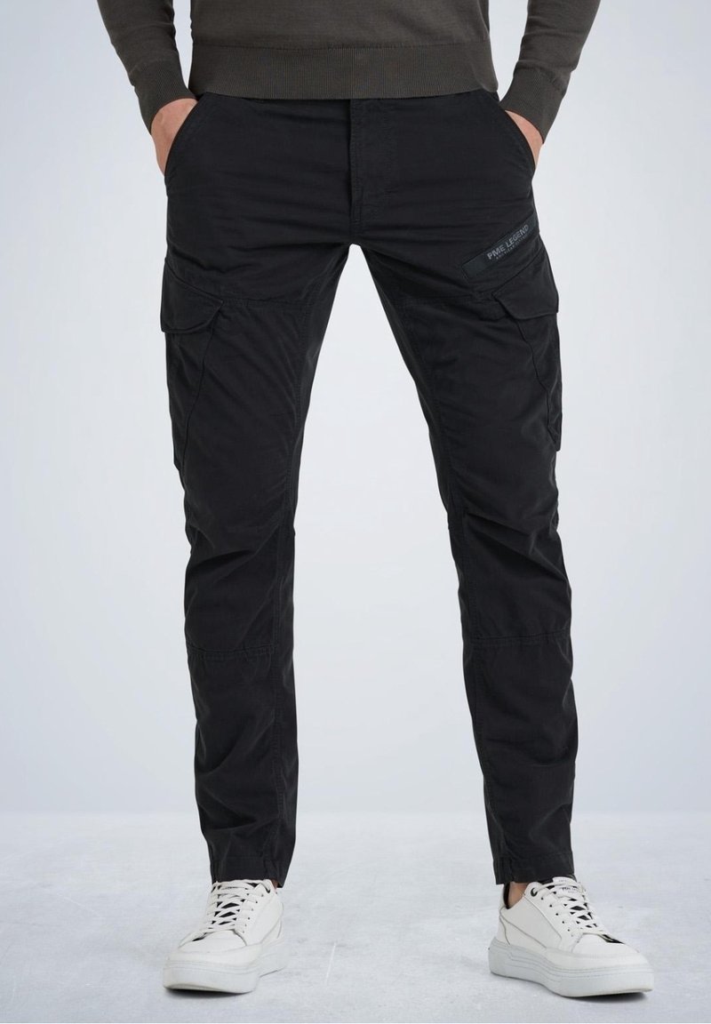 Homme debout les mains dans les poches, portant un pantalon cargo noir avec des poches à rabat et des baskets blanches sur un fond clair.