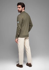Felpa verde oliva con scollatura rotonda e polsini a coste, abbinata a pantaloni color crema e mocassini marrone scuro.