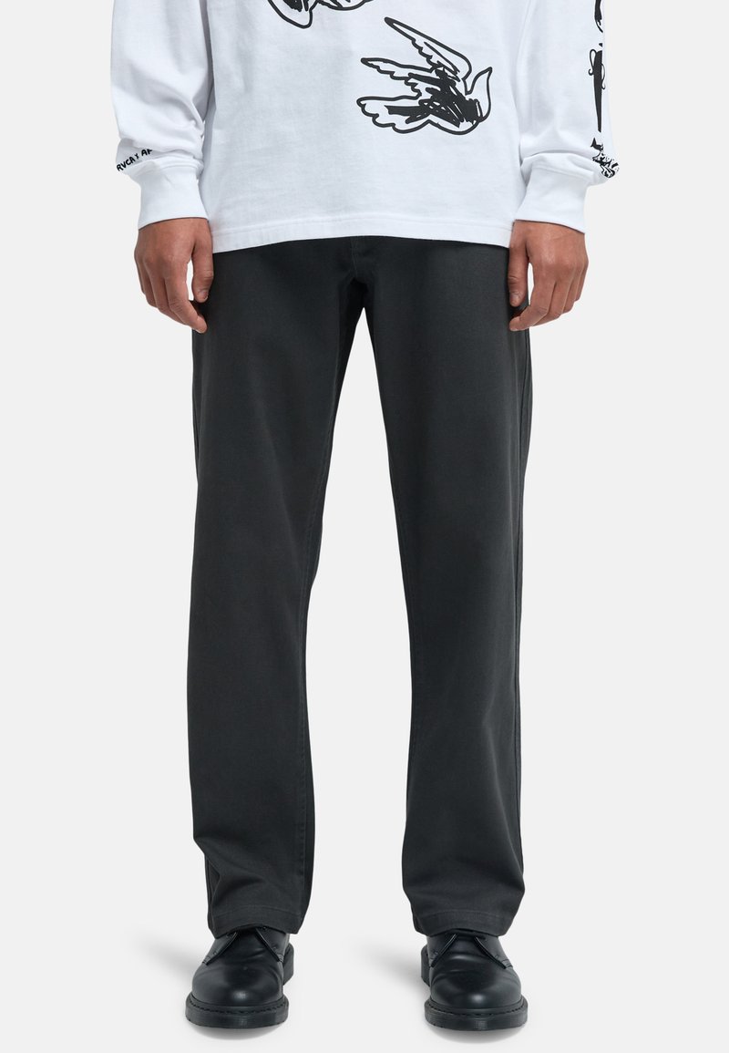 Onderste helft van een persoon met zwarte rechte broek, zwarte schoenen en een wit shirt met lange mouwen en zwarte vogelprints.