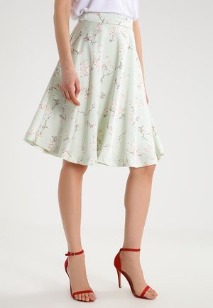 Kvinde iført en lysegrøn A-formet nederdel med lyserøde blomstergrene og røde ankelremssandaler med høje hæle, stående på en hvid baggrund.