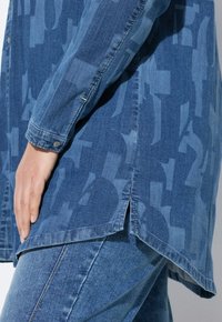 Camicia di jeans blu con un motivo sottile, maniche lunghe, bottone decorativo sul polsino, spacchi laterali e un design dell'orlo leggermente curvato.