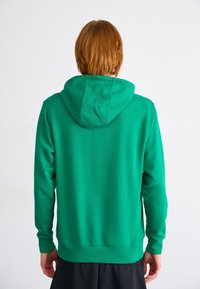 Person med kort röd hår, iklädd en grön hoodie och svarta shorts, står med ryggen vänd mot kameran mot en vit bakgrund.