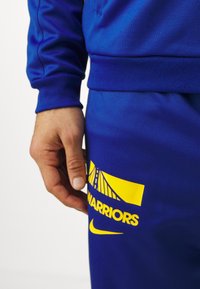 Mėlyna sportinė apranga, pagaminta iš lygaus audinio. Pasižymi ryškiu geltonu logotipu su užrašu "WARRIORS" ir Nike parašiniu swoosh ant kojos.