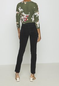 Kvinna stående med ryggen mot kameran, iklädd en olivgrön blommig långärmad överdel, svarta högmidjade byxor och vita klacksandaler mot en enfärgad bakgrund.