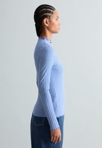 Femme aux cheveux tressés, vue de profil, portant un haut ajusté à manches longues rayé bleu et blanc ainsi qu'un jean en denim foncé, devant un fond uni.