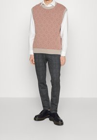 Pull en maille de couleur rouille et crème avec un motif en losanges, associé à un pantalon gris à carreaux et des chaussures noires avec des coutures jaunes.
