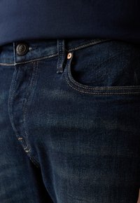 Jeans din denim albastru închis, cu un design clasic cu cinci buzunare, elemente de cupru și o ușoară decolorare. Vedere detaliată asupra centurii.