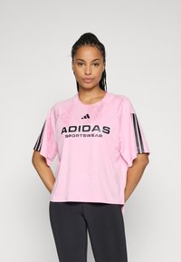 Kvinne iført rosa Adidas sportsklær skjorte med svart logo og striper på ermene, kombinert med svarte leggings, mot en ensfarget bakgrunn.