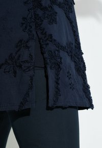 Donkerblauwe stof met getextureerde bloemrijke borduurwerk en een split, gelaagd over zwarte nauwsluitende broek.