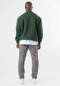 Uomo in piedi di spalle che indossa un maglione verde scuro, una maglietta bianca, pantaloni grigi e sneakers bianche su uno sfondo bianco.
