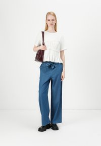Haut smocqué blanc avec ourlet volanté, associé à un pantalon en denim large bleu et des chaussures noires chunky. Sac à main bordeaux suspendu à l'épaule.