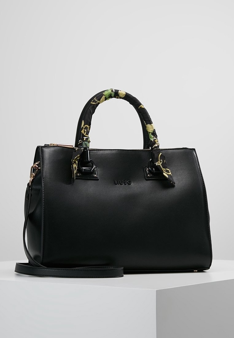 Sac à main structuré noir avec des poignées enveloppées de tissu à motifs floraux et une bandoulière amovible, posé sur une surface blanche.
