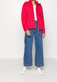 Rote gepolsterte Jacke mit Kapuze, mit einem Reißverschluss und horizontalen Nähten. Kombiniert mit weit geschnittenen blauen Denim-Jeans und weißen Sneakers.