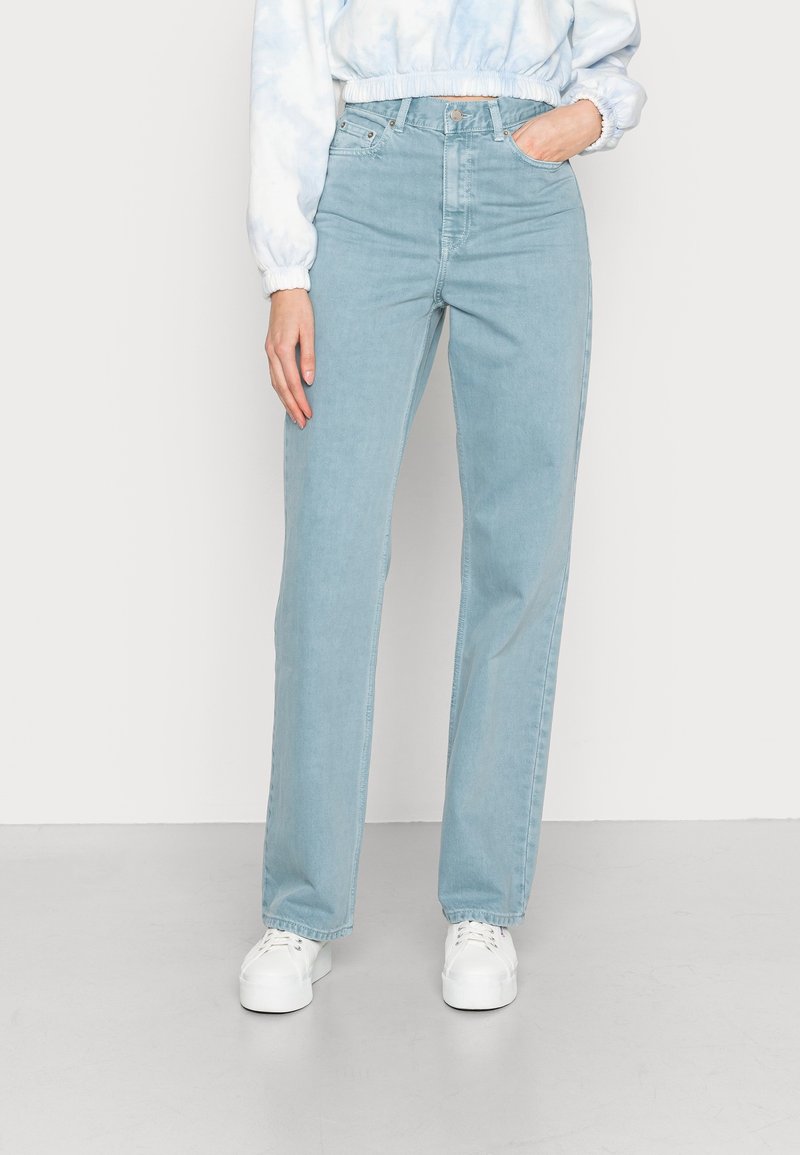 Femme portant un jean droit taille haute bleu clair, des baskets blanches à plateforme, et un sweat-shirt court bleu clair tie-dye, debout avec une main dans la poche.