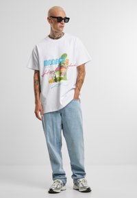 Biela bavlnená tričko s farebnou grafickou potlačou a textom "MONACO". Svetlomodré denimy a športové topánky dopĺňajú outfit.