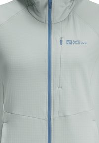 Giacca grigia chiara con motivo testurizzato, cerniera blu e logo. Presenta un colletto alto e una tasca sul petto con cerniera.