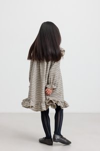 Une enfant aux longs cheveux noirs porte une robe à motifs et des collants noirs, se tenant de dos face au spectateur dans un espace minimaliste.
