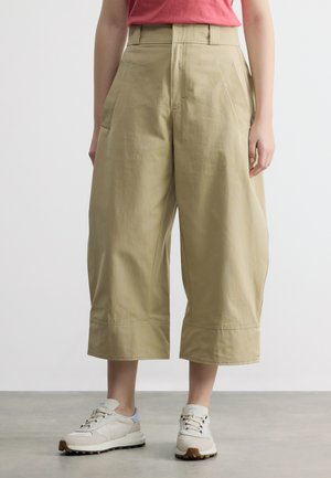 Άτομο που φοράει μπεζ φαρδύ cropped παντελόνι με μεγάλες πλαϊνές τσέπες, συνδυασμένο με λευκά και μπεζ αθλητικά παπούτσια και ένα κόκκινο τοπ.