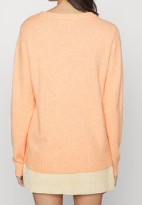 Suéter de punto naranja claro con cuello redondo, puños y dobladillo acanalados. Llevado sobre una falda de pana crema texturizada.