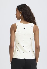 Donna con capelli lunghi e ricci che indossa una canottiera bianca con stampe di piccoli fiori neri, vista da dietro, con le mani nelle tasche dei pantaloni neri.