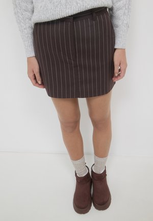 Eine Person trägt einen grauen Zopfmusterpullover, einen braun gestreiften Rock, beige Socken und dunkelbraune klobige Stiefeletten vor einem schlichten Hintergrund.