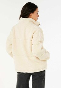 Crème-kleurige fleece jas met een hoge kraag, textuur, oversized pasvorm en stiksels, gedragen over donkere spijkerbroek.